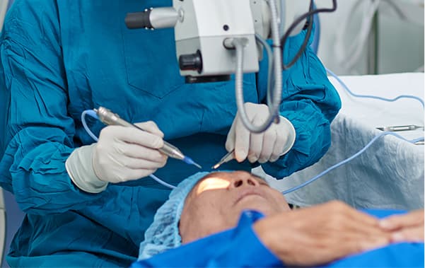 A surgeon in blue gown and gloves working under an operating microscope over a patient lying on the table