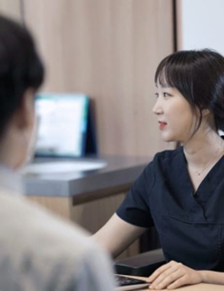 A doctor in dark scrubs seated at a desk in a bright consultation room, turned toward a patient who is seen from behind