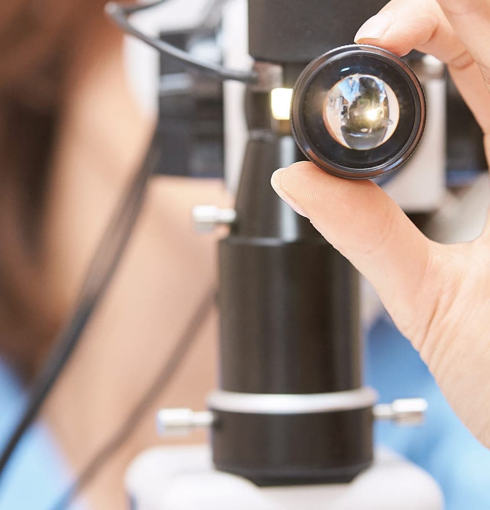 A hand holding a small examination lens in front of a slit-lamp microscope, with the examiner blurred in the background